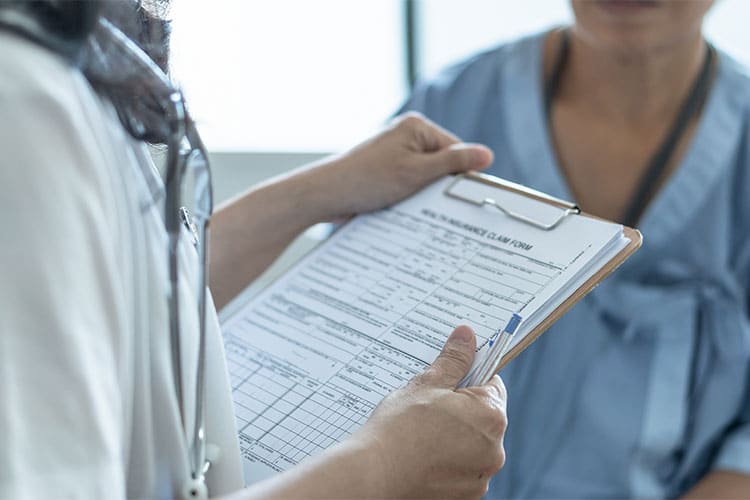 Un médico con un portapapeles hablando con un paciente. ¿Cuál es la diferencia entre la protección contra lesiones personales y la cobertura de pagos médicos al presentar una reclamación por lesiones personales?