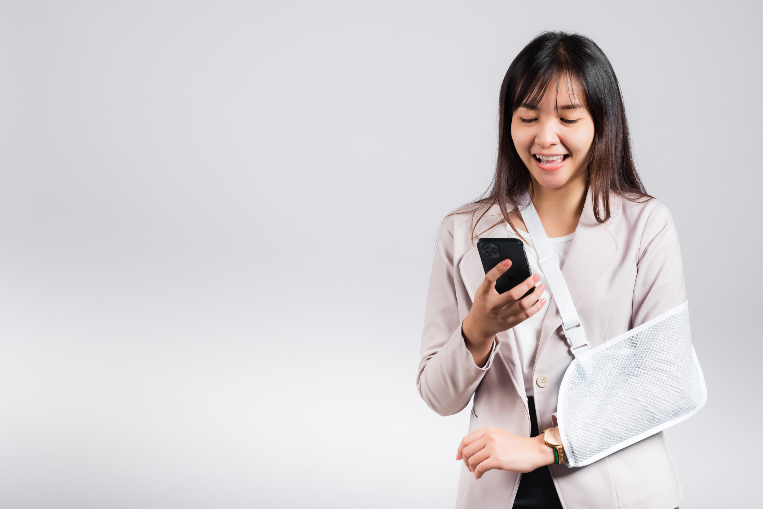 Woman confident smiling broken arm after accident and wear arm splint for treatment and hold smartphone, happy Asian female sling supported hand isolated on white background, social media, copy space. Social Media and Personal Injury Claims