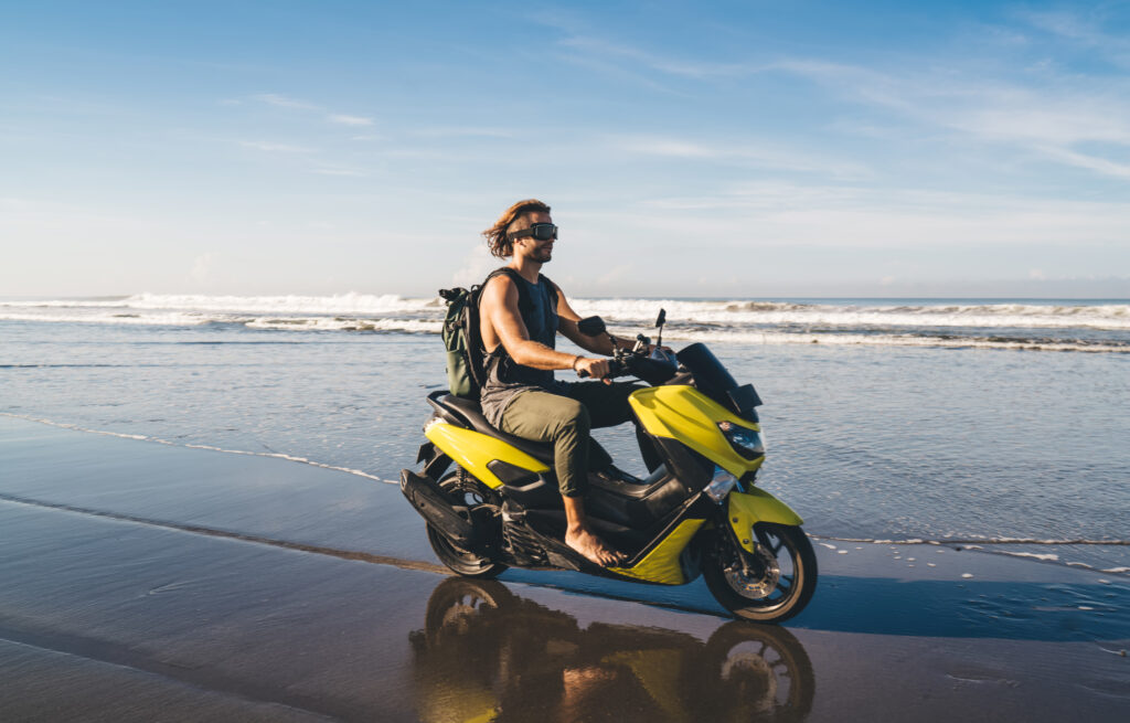 Longitud total de un viajero masculino con gafas conduciendo una motocicleta sobre arena mojada cerca del mar ondeante mientras mira hacia otro lado y descansa durante las vacaciones.