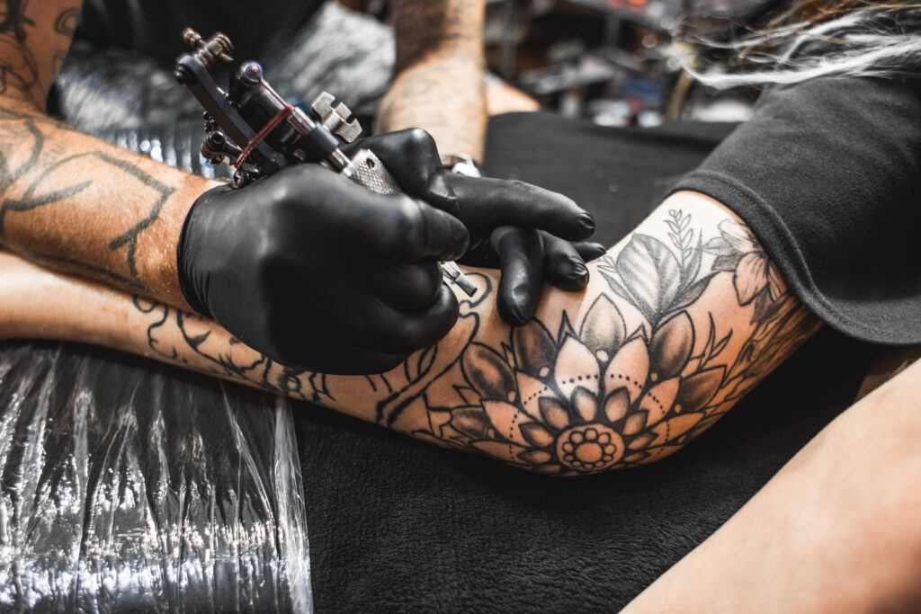 Hazte un tatuaje: una chica con rastas en un salón de tatuajes. El maestro crea una imagen en el cuerpo de una joven hermosa. Primer plano de las manos y la máquina de tatuar. ¿Qué edad hay que tener para hacerse un tatuaje?