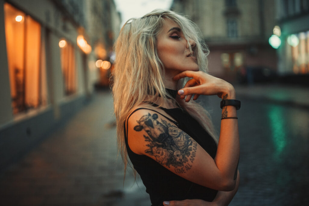 Retrato de una joven con un tatuaje en el hombro de pie en una calle de la ciudad al anochecer. Al fondo, se ven las luces de la ciudad. ¿Qué edad hay que tener para hacerse un tatuaje en Estados Unidos?