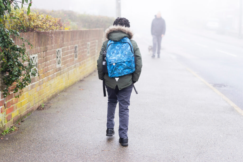 Niño caminando hacia la escuela solo en la acera en la niebla con una mochila puesta.